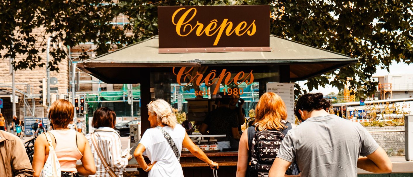 Menschen stehen an einem Crêpes-Stand vor dem Stuttgarter Hauptbahnhof. Der Stand ist mit einem Schild 'Crêpes seit 1980' versehen., © SMG, Sarah Schmid