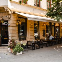 Ein Café an einer Straßenecke mit Außenbestuhlung, Pflanzen und hängenden Lampen. Menschen sitzen an Tischen unter einem Sonnenschutz., © SMG, Sarah Schmid Ein Café an einer Straßenecke mit Außenbestuhlung, Pflanzen und hängenden Lampen. Menschen sitzen an Tischen unter einem Sonnenschutz., © SMG, Sarah Schmid