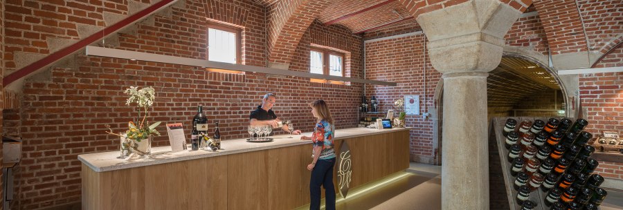 A bar in a wine cellar with brick walls. A man pours wine for a woman. Wine bottles are displayed on the counter., © Collegium Wirtemberg