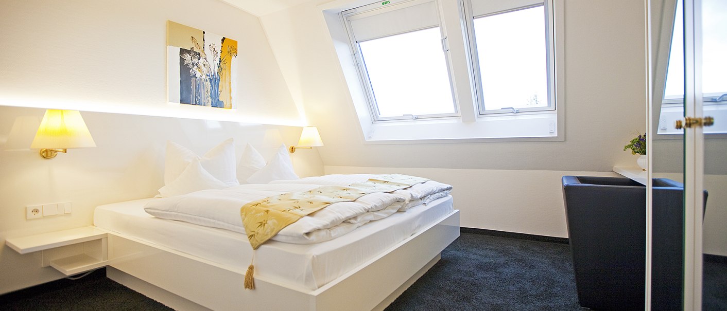 Helles Schlafzimmer mit Doppelbett, zwei Dachfenstern, Wandkunst und Nachttischlampen. Der Raum wirkt modern und einladend., &copy; Hotel Krone