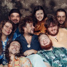 Acht fr&ouml;hliche Personen posieren eng beieinander und l&auml;cheln in die Kamera. Sie tragen bunte, l&auml;ssige Kleidung vor einem dunklen Hintergrund., &copy; links im Bild