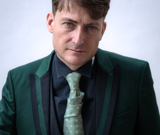 Ein Mann trägt einen eleganten grünen Anzug mit passender Krawatte und schaut direkt in die Kamera. Der Hintergrund ist schlicht und hell., © Rosenau Kultur e.V. Ein Mann trägt einen eleganten grünen Anzug mit passender Krawatte und schaut direkt in die Kamera. Der Hintergrund ist schlicht und hell., © Rosenau Kultur e.V.