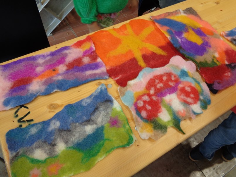 Bunte Filzarbeiten auf einem Holztisch. Zu sehen sind Landschaften und Pilze in leuchtenden Farben. Eine Person in grünem Pullover ist teilweise sichtbar., © KVWH Bunte Filzarbeiten auf einem Holztisch. Zu sehen sind Landschaften und Pilze in leuchtenden Farben. Eine Person in grünem Pullover ist teilweise sichtbar., © KVWH