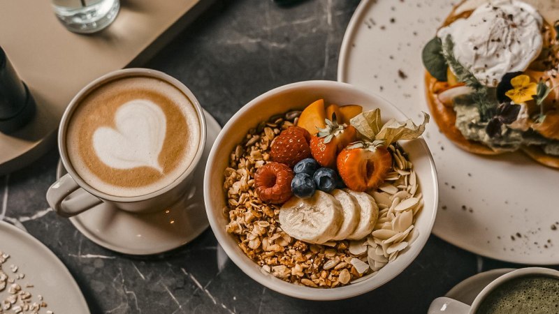 Ein Frühstückstisch mit einer Tasse Kaffee mit Herz-Latte-Art, einer Schüssel Müsli mit Früchten und einem Teller mit Waffeln und Avocado., © Fritz Stuttgart Ein Frühstückstisch mit einer Tasse Kaffee mit Herz-Latte-Art, einer Schüssel Müsli mit Früchten und einem Teller mit Waffeln und Avocado., © Fritz Stuttgart