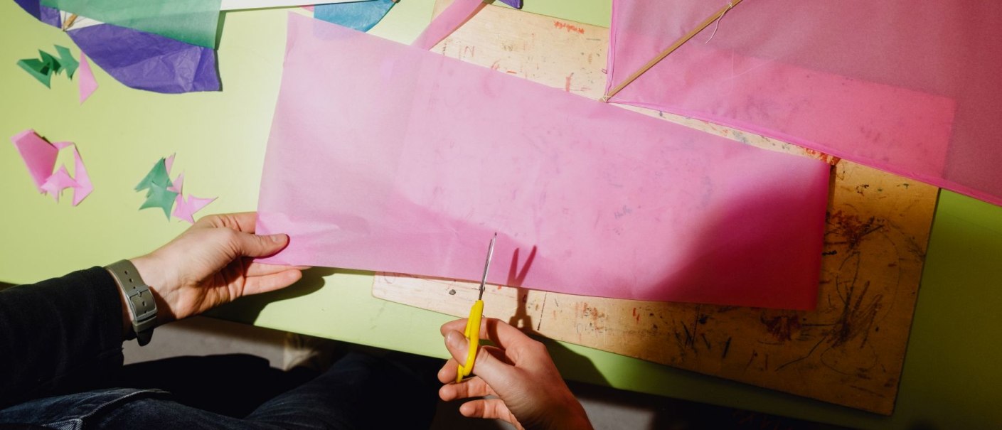 Eine Person schneidet rosa Papier mit einer gelben Schere. Auf dem Tisch liegen bunte Papierst&uuml;cke und ein Holzbrett., &copy; Linden-Museum Stuttgart, Foto: Harald V&ouml;lkl