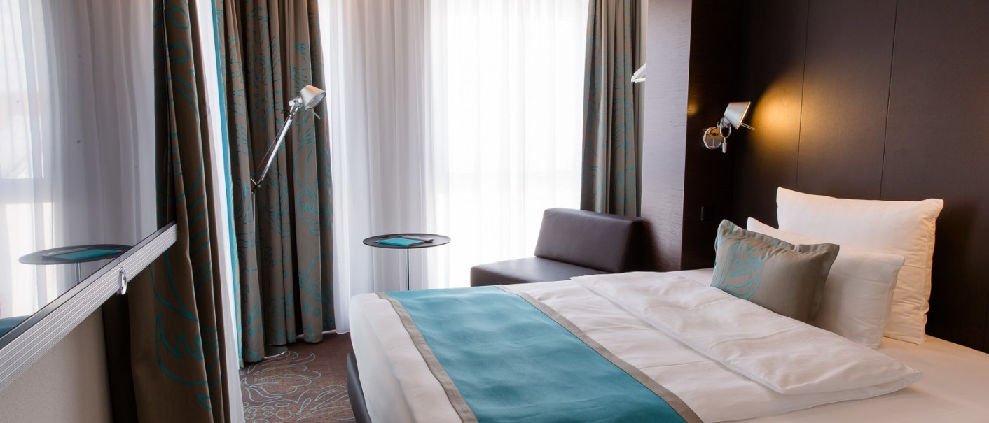 Modern hotel room with double bed, blue accents, sofa, floor lamp and large windows with curtains., &copy; Motel One Stuttgart-Bad Cannstatt