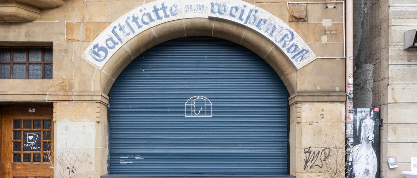 Entrance with the inscription 'Gaststätte zum weißen Roß' above a blue roller shutter. Next to it is a wooden door with a 'Card Only' sign., © Stuttgart-Marketing GmbH, Sarah Schmid Entrance with the inscription 'Gaststätte zum weißen Roß' above a blue roller shutter. Next to it is a wooden door with a 'Card Only' sign., © Stuttgart-Marketing GmbH, Sarah Schmid