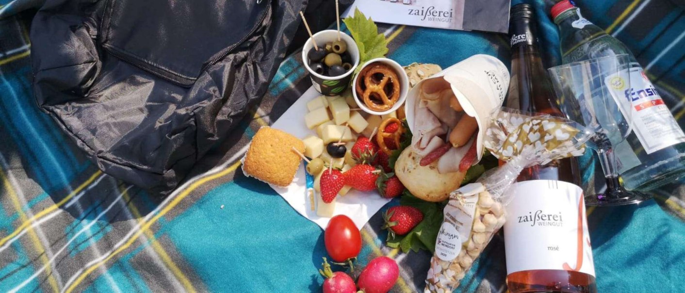 A picnic on a checkered blanket with cheese, olives, strawberries, bread, wine and a bottle of water. A rucksack lies next to it., © Weingut Zaißerei