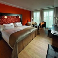 Modern hotel room with large bed, orange wall, sofa, desk and TV. Light curtains and wooden floor create a cozy atmosphere., &copy; Parkhotel Stuttgart Messe-Airport