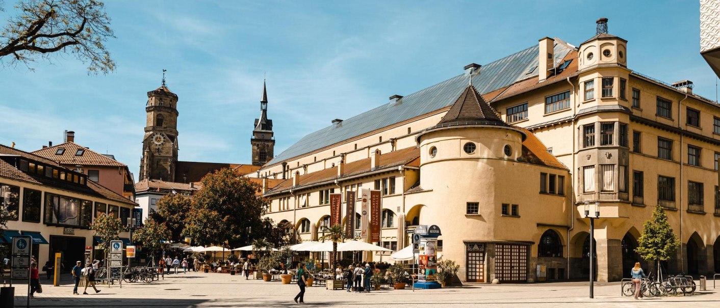 Die Markthalle Stuttgart bei sonnigem Wetter, umgeben von Menschen, Fahrr&auml;dern und B&auml;umen. Historische Geb&auml;ude im Hintergrund., &copy; Stuttgart Marketing GmbH, Sarah Schmid
