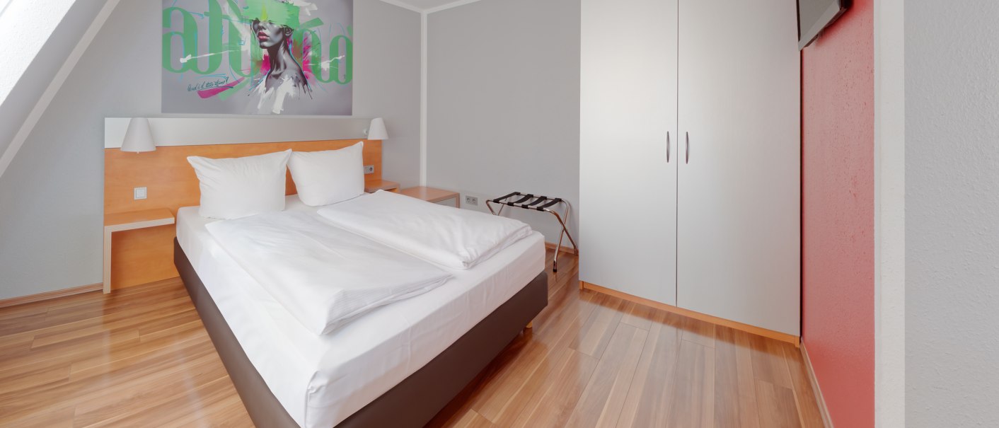 Modernes Hotelzimmer mit Doppelbett, weißem Kleiderschrank, Wandkunst und Flachbildfernseher. Holzboden und rote Akzentwand., © attimo Hotel Stuttgart Modernes Hotelzimmer mit Doppelbett, weißem Kleiderschrank, Wandkunst und Flachbildfernseher. Holzboden und rote Akzentwand., © attimo Hotel Stuttgart
