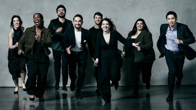Acht Personen in eleganter Kleidung laufen lächelnd auf die Kamera zu. Der Hintergrund ist eine graue Wand, der Boden reflektiert leicht., © Württembergische Staatstheater Stuttgart Acht Personen in eleganter Kleidung laufen lächelnd auf die Kamera zu. Der Hintergrund ist eine graue Wand, der Boden reflektiert leicht., © Württembergische Staatstheater Stuttgart