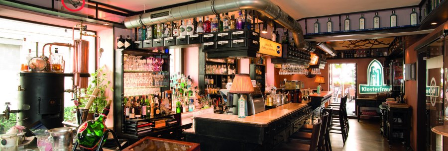 Bar mit Flaschen, Gl&auml;sern und Kupferdestille. Klosterfrau-Logo leuchtet im Hintergrund. Gem&uuml;tliche Atmosph&auml;re mit Holzm&ouml;beln und Lampen., &copy; Buschwerk Mediendesign-Wilhelmer Gastronomie
