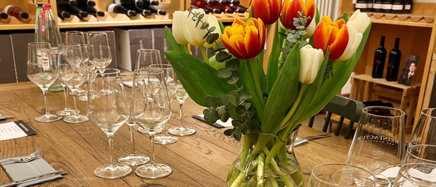 Ein Tisch mit Weingläsern und einem Tulpenstrauß, umgeben von Weinflaschen in Regalen. Elegante Atmosphäre für einen Weinabend., © Weinfactum Bad Cannstatt Ein Tisch mit Weingläsern und einem Tulpenstrauß, umgeben von Weinflaschen in Regalen. Elegante Atmosphäre für einen Weinabend., © Weinfactum Bad Cannstatt