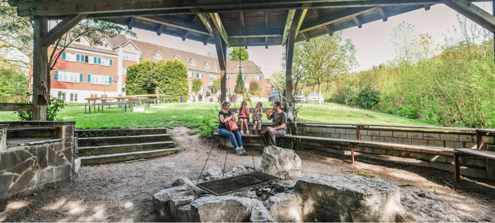 &Uuml;berdachter Grillplatz mit Sitzb&auml;nken und Personen, im Hintergrund ein Geb&auml;ude mit blauen Fensterl&auml;den., &copy; DJH Baden-W&uuml;rttemberg e.V.