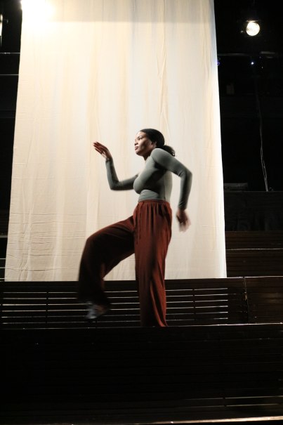 A person is dancing on a stage in front of a large, light-colored curtain. She is wearing a gray top and brown pants., &copy; Amina Ousman