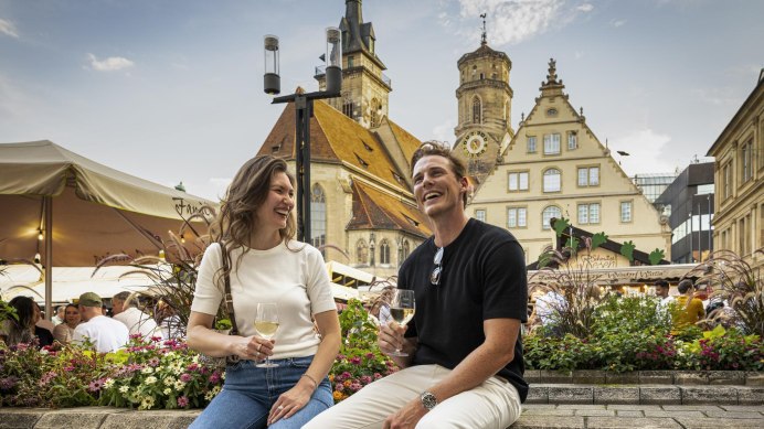 Ein Paar genießt das Stuttgarter Weindorf auf dem Schillerplatz, umgeben von Blumen und historischen Gebäuden. Beide halten Weingläser und lachen., © Stuttgart-Marketing GmbH, Sarah Schmid