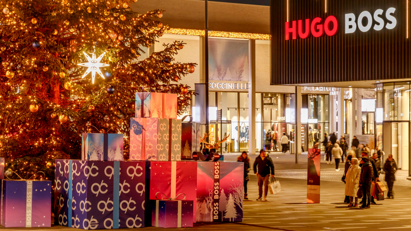 Weihnachtlich geschmückter Platz mit großem Baum und Geschenken vor dem Hugo Boss Geschäft. Menschen flanieren in der festlichen Atmosphäre., © OUTLETCITY AG Weihnachtlich geschmückter Platz mit großem Baum und Geschenken vor dem Hugo Boss Geschäft. Menschen flanieren in der festlichen Atmosphäre., © OUTLETCITY AG