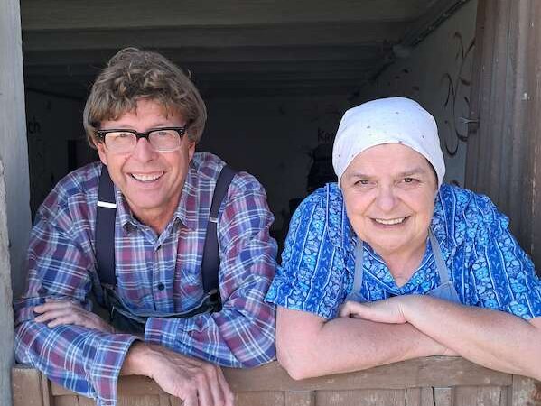 Zwei Personen lächeln über eine Holztür. Eine trägt ein kariertes Hemd, die andere ein blaues Kopftuch. Sie wirken freundlich und entspannt., © SBEntertainment GmbH & Co. KG Zwei Personen lächeln über eine Holztür. Eine trägt ein kariertes Hemd, die andere ein blaues Kopftuch. Sie wirken freundlich und entspannt., © SBEntertainment GmbH & Co. KG