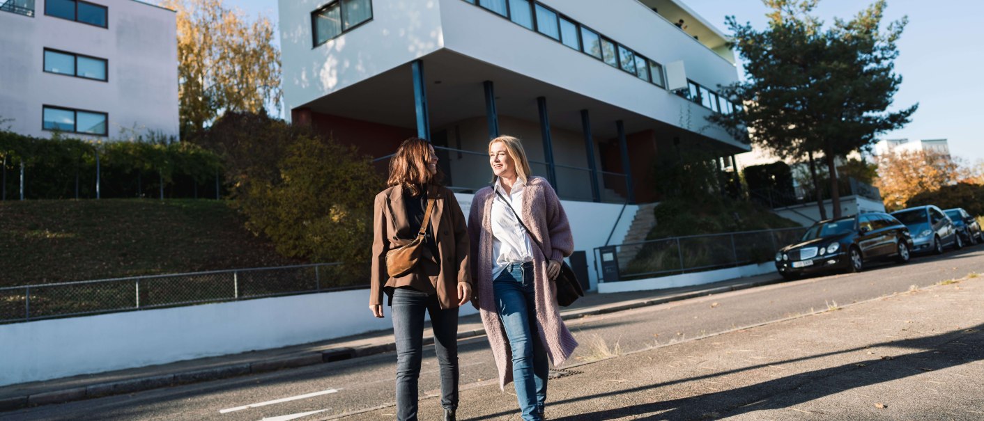 Two women walk in front of the Weissenhof Museum in Stuttgart. The building by Le Corbusier is modern and white, with clear lines and large windows., © SMG, WP Steinheisser Two women walk in front of the Weissenhof Museum in Stuttgart. The building by Le Corbusier is modern and white, with clear lines and large windows., © SMG, WP Steinheisser