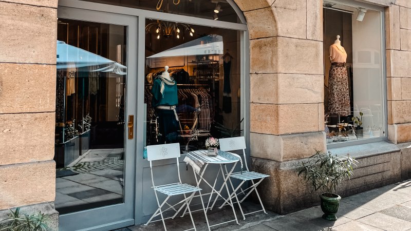 A small store with a display window, decorated with clothes. There are two white chairs and a table on the sidewalk in front of it., &copy; MAGASIN Gei&szlig;strasse 15, Stuttgart