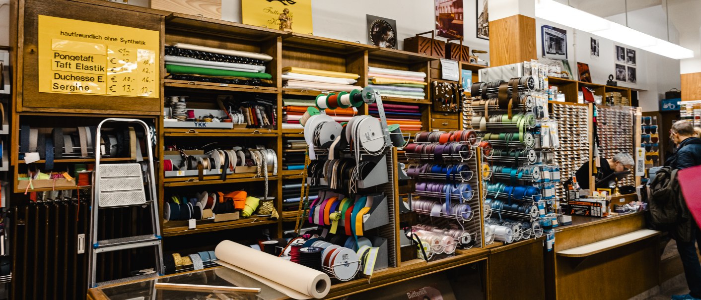 Ein Stoffladen mit Regalen voller bunter Bänder und Stoffrollen. Eine Leiter steht an der Wand, und ein Schild weist auf hautfreundliche Stoffe hin., © SMG Stuttgart Marketing GmbH - Sarah Schmid