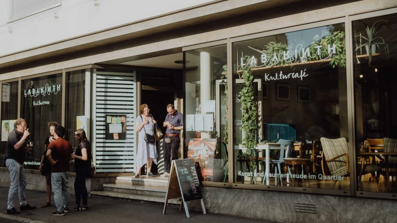 People chat in front of the LABYRINTH cultural center & café. The windows are decorated with plants and there is a sign in front of the entrance., © Christoph Steinweg
