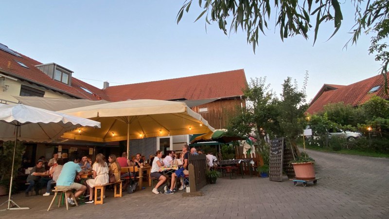 Menschen sitzen unter großen Sonnenschirmen in einem gemütlichen Hof mit roten Dächern und Bäumen., © Weinhof Zaiß Menschen sitzen unter großen Sonnenschirmen in einem gemütlichen Hof mit roten Dächern und Bäumen., © Weinhof Zaiß