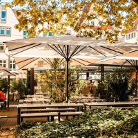 Sonniger Café-Außenbereich mit weißen Sonnenschirmen, Holztischen und Pflanzen. Menschen sitzen entspannt unter den Schirmen. Gebäude im Hintergrund., © Stuttgart Marketing GmbH, Sarah Schmid Sonniger Café-Außenbereich mit weißen Sonnenschirmen, Holztischen und Pflanzen. Menschen sitzen entspannt unter den Schirmen. Gebäude im Hintergrund., © Stuttgart Marketing GmbH, Sarah Schmid