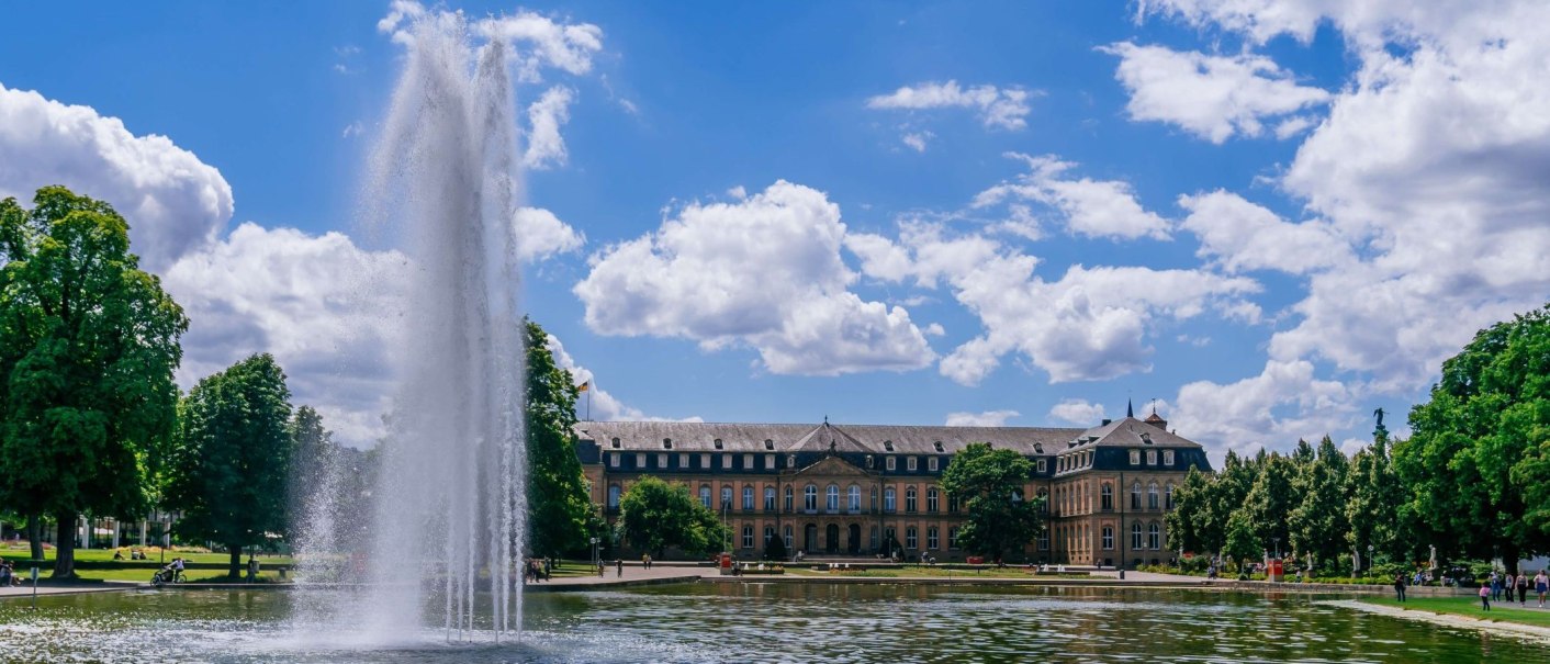 Das Bild zeigt das Neue Schloss in Stuttgart mit einem großen Springbrunnen im Vordergrund und dem Eckensee. Der Himmel ist blau mit weißen Wolken., © Thomas Niedermüller