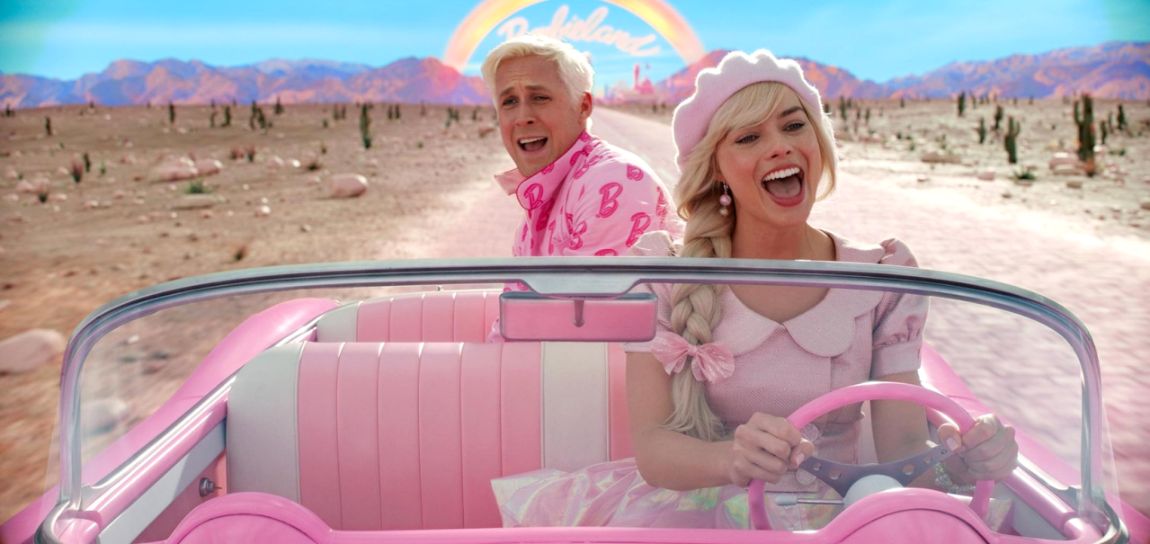 Zwei Personen in einem pinken Cabrio fahren durch eine Wüstenlandschaft. Ein Regenbogen und der Schriftzug "Barbie" sind im Hintergrund zu sehen., © Warner Bros.