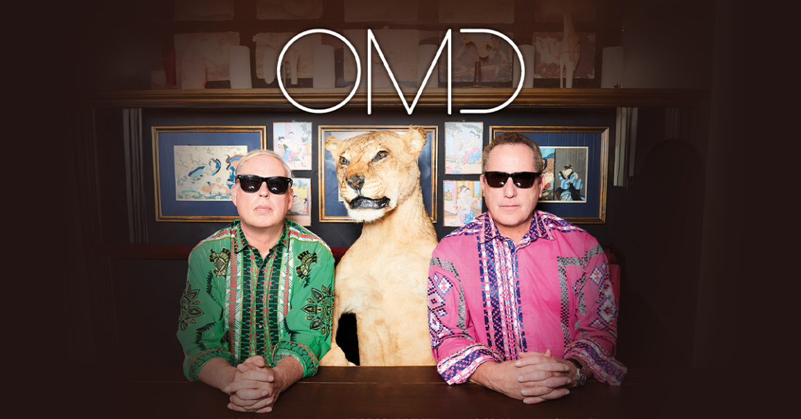 Zwei M&auml;nner in bunten Hemden und Sonnenbrillen sitzen neben einem ausgestopften L&ouml;wenkopf. Im Hintergrund sind gerahmte Bilder zu sehen. Oben steht 'OMD'., &copy; links im Bild