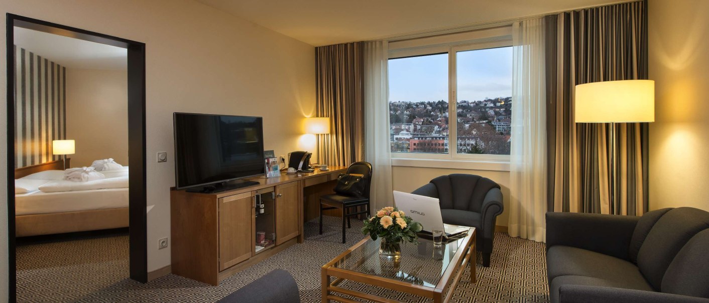 Elegante Junior Suite mit Wohnzimmer, Sofa, Tisch, Fernseher und großem Fenster mit Stadtblick. Schlafzimmer im Hintergrund sichtbar., © Maritim Hotelgesellschaft mbH Elegante Junior Suite mit Wohnzimmer, Sofa, Tisch, Fernseher und großem Fenster mit Stadtblick. Schlafzimmer im Hintergrund sichtbar., © Maritim Hotelgesellschaft mbH