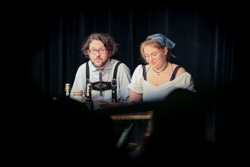 Zwei Personen in traditioneller Kleidung sitzen auf einer Bühne mit Mikrofonen. Der Mann trägt eine Brille und Hosenträger, die Frau ein Kopftuch., © Theaterhaus Stuttgart e.V.