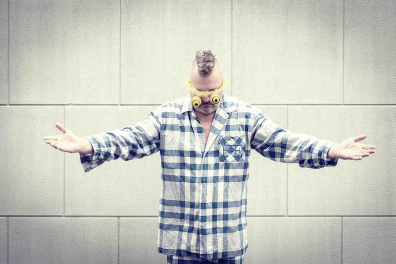 Eine Person in einem blau-weiß karierten Pyjama trägt gelbe Brillen und steht mit ausgestreckten Armen vor einer grauen Wand., © Rosenau Kultur e.V. Eine Person in einem blau-weiß karierten Pyjama trägt gelbe Brillen und steht mit ausgestreckten Armen vor einer grauen Wand., © Rosenau Kultur e.V.