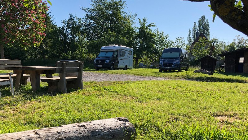 Wohnmobilstellplatz mit zwei Wohnmobilen, Holztisch und Bänken unter einem blühenden Baum. Im Hintergrund sind kleine Holzhütten zu sehen., © Bio Weingut Zimmer Wohnmobilstellplatz mit zwei Wohnmobilen, Holztisch und Bänken unter einem blühenden Baum. Im Hintergrund sind kleine Holzhütten zu sehen., © Bio Weingut Zimmer