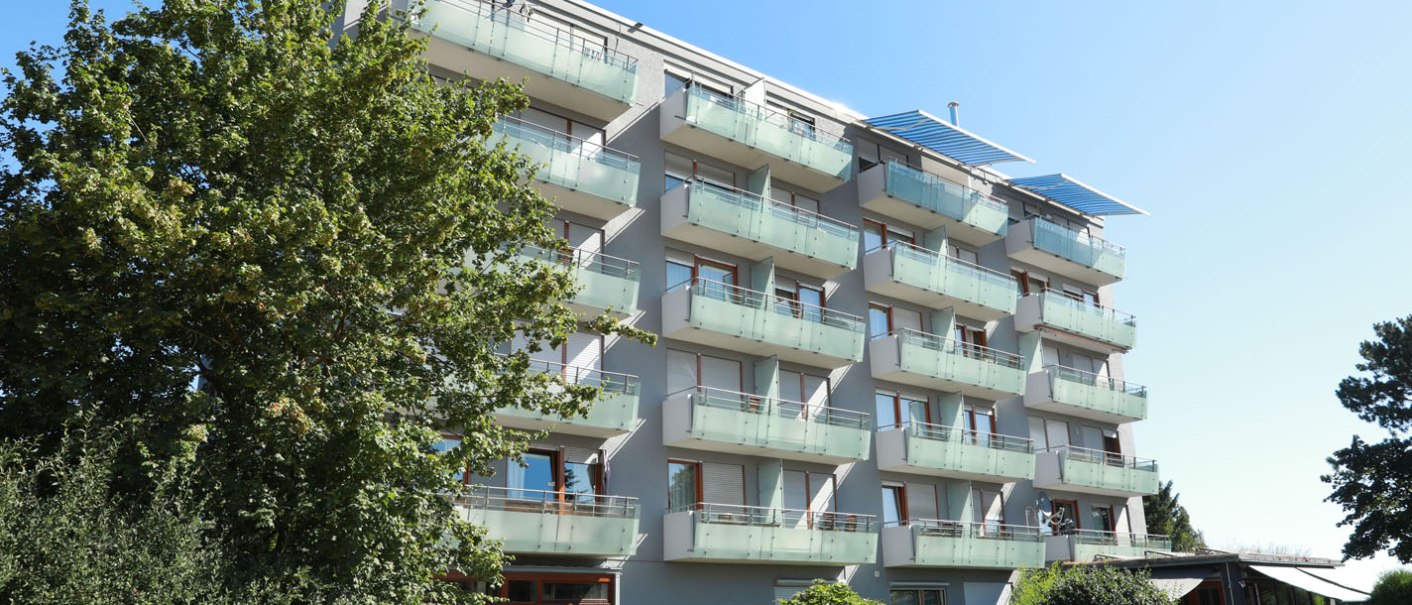 A modern, multi-storey residential building with balconies and glass railings, surrounded by trees and a clear blue sky., © TOMAS