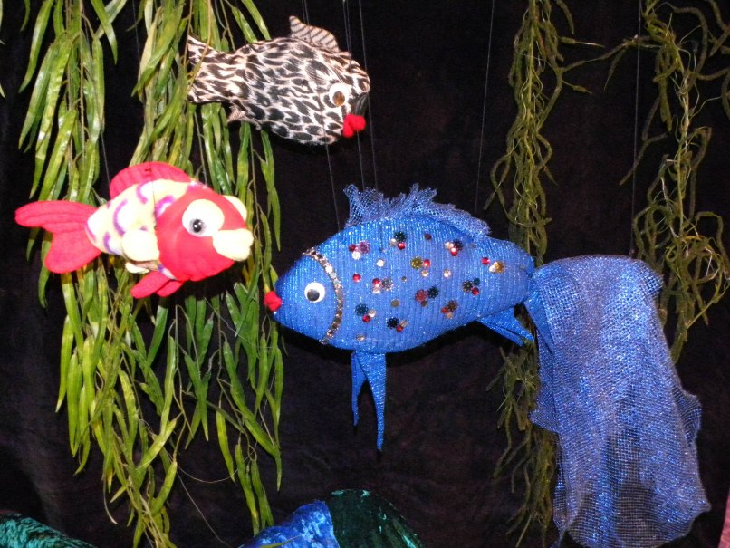 Drei bunte Stofffische, darunter ein blauer mit Glitzer, hängen vor einem dunklen Hintergrund mit grünen Pflanzen., © Nellys Puppentheater Drei bunte Stofffische, darunter ein blauer mit Glitzer, hängen vor einem dunklen Hintergrund mit grünen Pflanzen., © Nellys Puppentheater