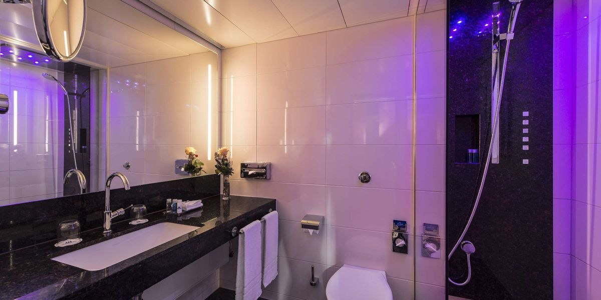 Modernes Badezimmer mit dunklem Waschtisch, großem Spiegel, Dusche und lila Beleuchtung. Handtücher und Blumen dekorieren den Raum., © Maritim Hotelgesellschaft mbH Modernes Badezimmer mit dunklem Waschtisch, großem Spiegel, Dusche und lila Beleuchtung. Handtücher und Blumen dekorieren den Raum., © Maritim Hotelgesellschaft mbH