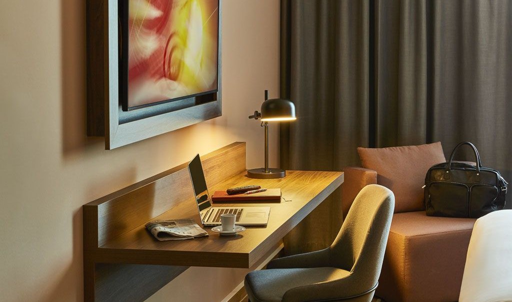 Modernes Hotelzimmer mit Schreibtisch, Laptop, Lampe und Sessel. Ein Kunstwerk hängt an der Wand, eine Tasche steht auf dem Sofa., © Pullman Stuttgart Fontana