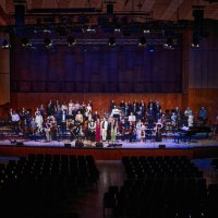 Ein Orchester mit Chor steht auf einer B&uuml;hne in einem Konzertsaal. Die Musiker tragen festliche Kleidung, der Saal ist mit leeren Stuhlreihen gef&uuml;llt., &copy; Marc Sch&auml;fer