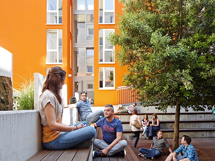Menschen entspannen in einem modernen Innenhof mit orangen Wänden und einem Baum. Sie sitzen auf Holzbänken und unterhalten sich., © Studierendenwerk Stuttgart
