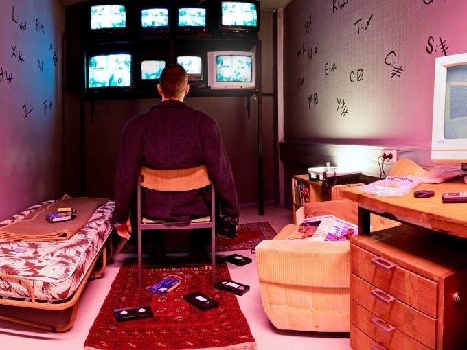Ein Mann sitzt in einem Raum mit Monitoren, VHS-Kassetten und kryptischen Symbolen an den Wänden. Der Raum wirkt überwacht und geheimnisvoll., © ExitGames Stuttgart Ein Mann sitzt in einem Raum mit Monitoren, VHS-Kassetten und kryptischen Symbolen an den Wänden. Der Raum wirkt überwacht und geheimnisvoll., © ExitGames Stuttgart