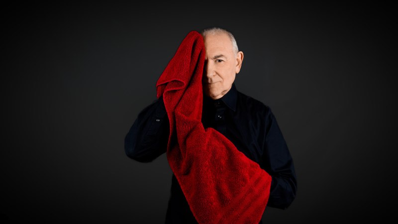 Ein Mann in dunkler Kleidung hält ein rotes Handtuch vor sein Gesicht, vor einem dunklen Hintergrund., © Carlos Martinez Ein Mann in dunkler Kleidung hält ein rotes Handtuch vor sein Gesicht, vor einem dunklen Hintergrund., © Carlos Martinez