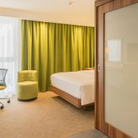 Modernes Hotelzimmer mit Doppelbett, grünem Sessel, Schreibtisch und Blick ins Badezimmer mit Dusche. Helle Vorhänge und grüne Gardinen., © Hampton by Hilton Stuttgart City Centre