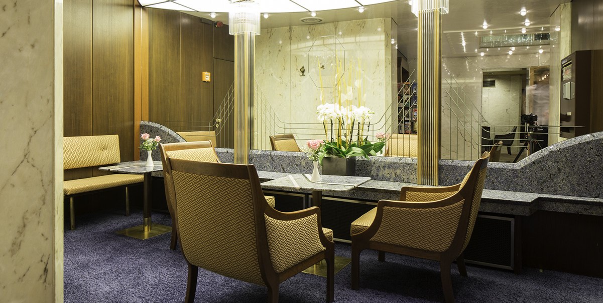 Elegante Sitzecke mit gepolsterten Sesseln, einem Tisch und Blumenarrangement. Holzvertäfelung und moderne Beleuchtung schaffen eine stilvolle Atmosphäre., © Hotel Royal