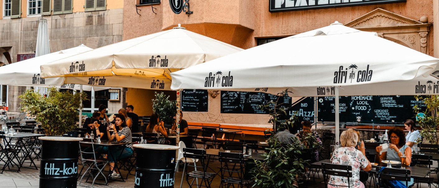 Außenbereich des Cafés Platzhirsch mit weißen Sonnenschirmen und Gästen an Tischen. Die Fassade ist orange, und es gibt Pflanzen als Dekoration., © SMG Stuttgart Marketing GmbH - Sarah Schmid