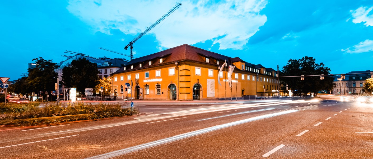 Gelbes Geb&auml;ude bei Abendd&auml;mmerung, beleuchtet von Stra&szlig;enlichtern. Autos ziehen Lichtspuren auf der Stra&szlig;e, Baukr&auml;ne im Hintergrund., &copy; Luca Siermann, ifa Galerie Stuttgart