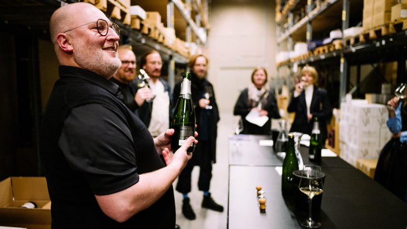 A group of people at a wine tasting in a storage room. One man is holding a wine bottle, while other participants are holding glasses., © Bernhard Kahrmann