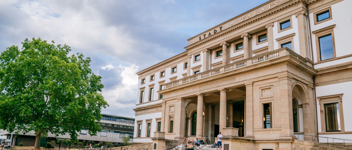Das StadtPalais in Stuttgart zeigt eine klassizistische Fassade mit Säulen. Ein großer Baum steht links, Menschen sitzen auf den Stufen., © SMG Thomas Niedermüller